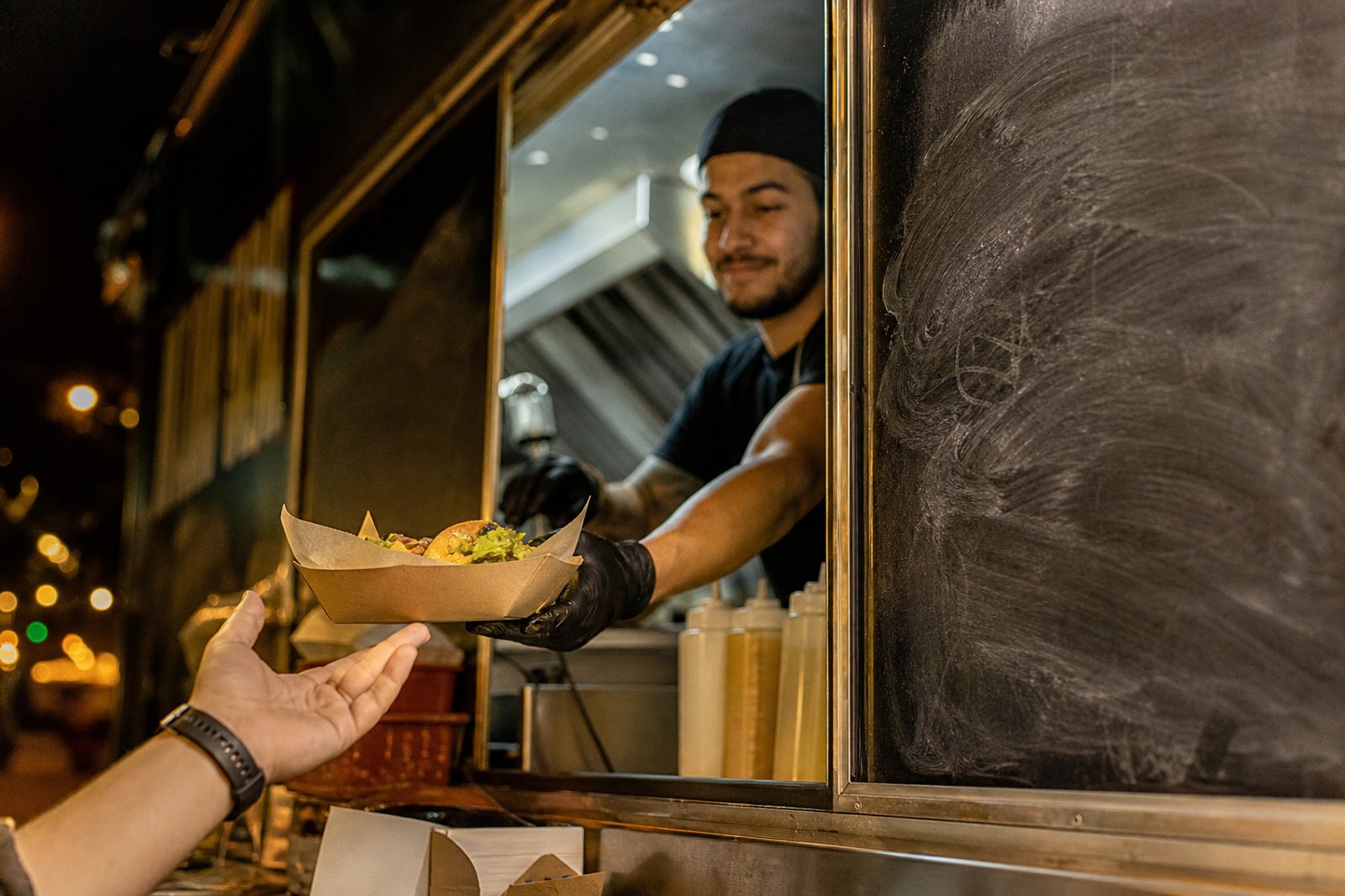 A cook handing a paper tray through a food truck service window beside a chalkboard menu under evening street lights
